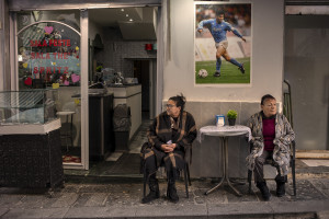 Two women are seen outside a bar near a poster depicting the Argentine soccer legend Diego Armando Maradona in Forcella street in Naples, Southern Italy on November 23, 2021. Diego Armando Maradona died on November 25 of last year and next November 25, 2021 will be the anniversary of his death.