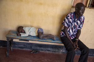 A child probably suffering from malaria sleeps on a bench, while a man waits his turn inside the Kalokol Gok Health Centre, located in Turkana County, northwest Kenya on October 16, 2024. Children often go fetching water with their mothers in areas where stagnant pools of water form and mosquitoes that carry the malaria parasite proliferate. Over the past three years Kenya has suffered from intense drought, partly caused by global warming, which also provides favourable conditions for the development of these mosquitoes.